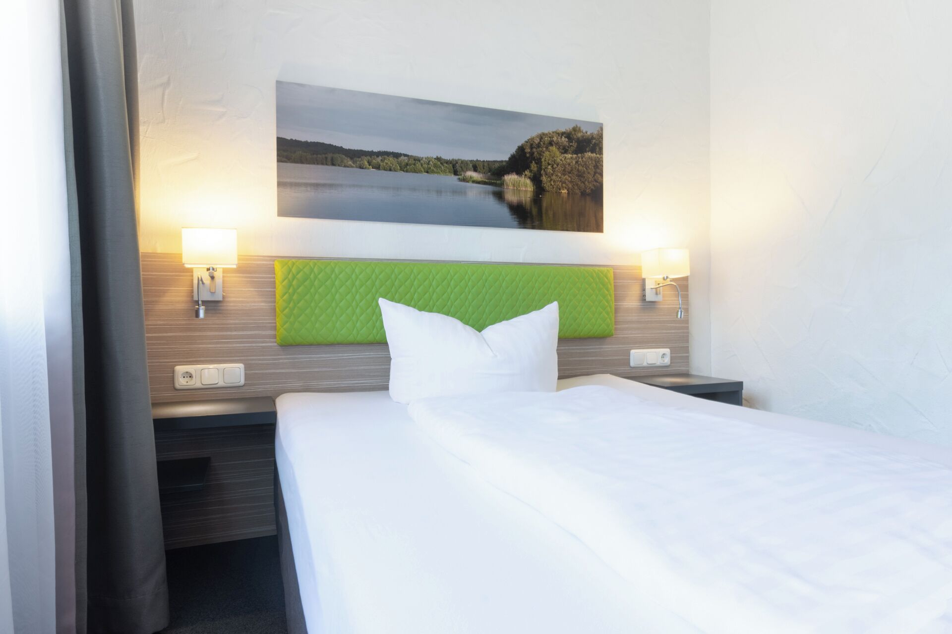 A hotel room with a bed, two bedside lamps, a green headboard, and a framed picture of a lake.
