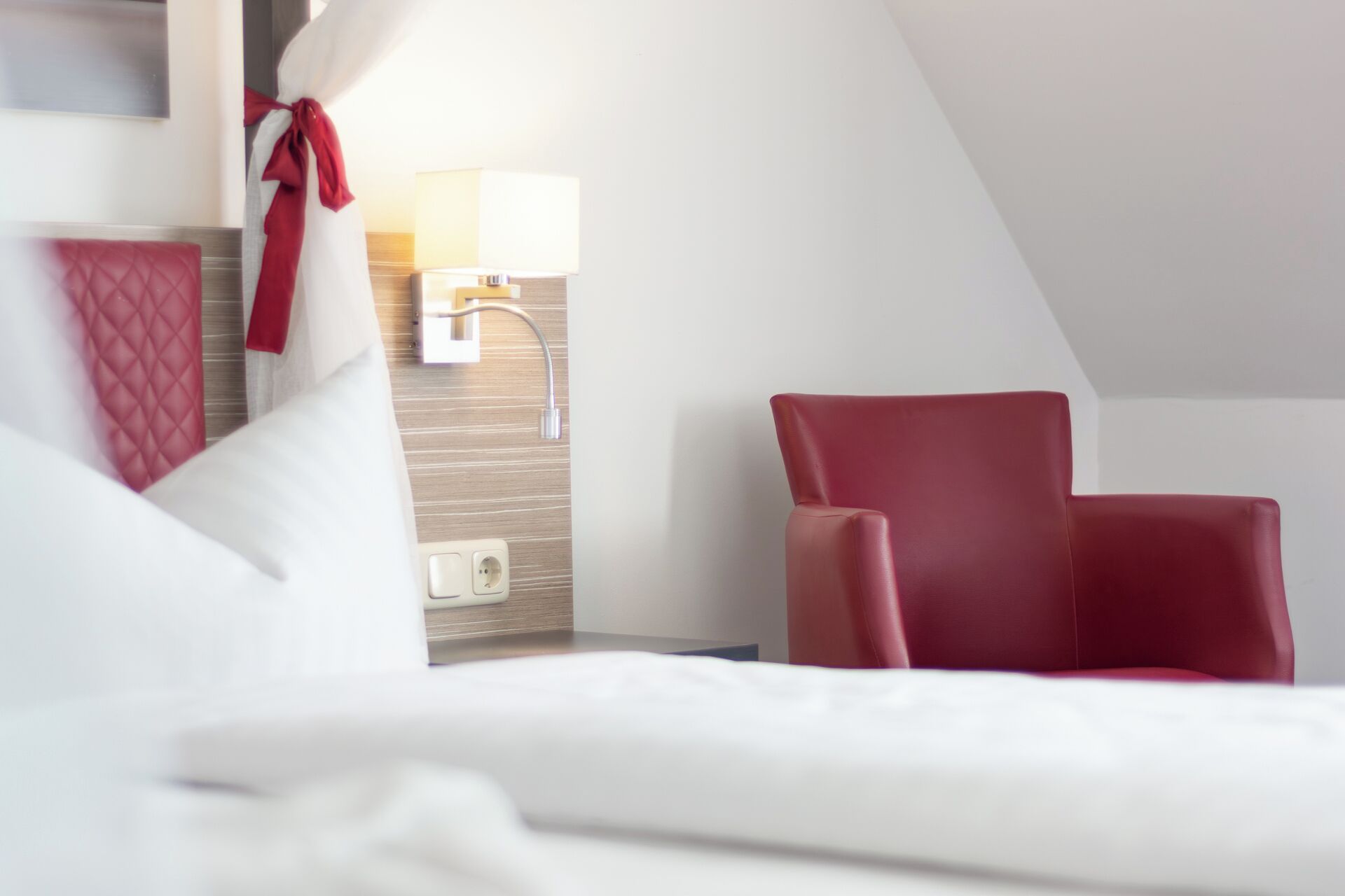 Una habitación de hotel con una cama, una silla roja, una lámpara y un enchufe de pared.