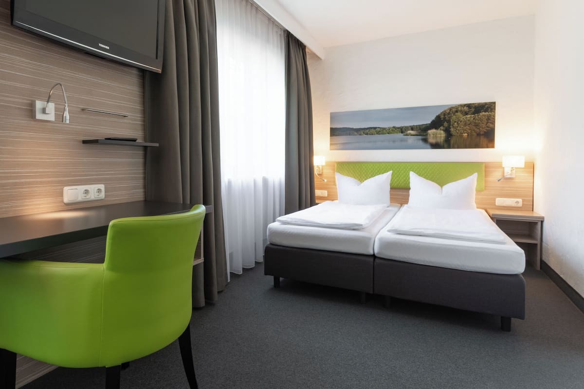 Une chambre d’hôtel avec un lit, un bureau et une chaise. Il y a un tableau au mur et une fenêtre avec des rideaux.