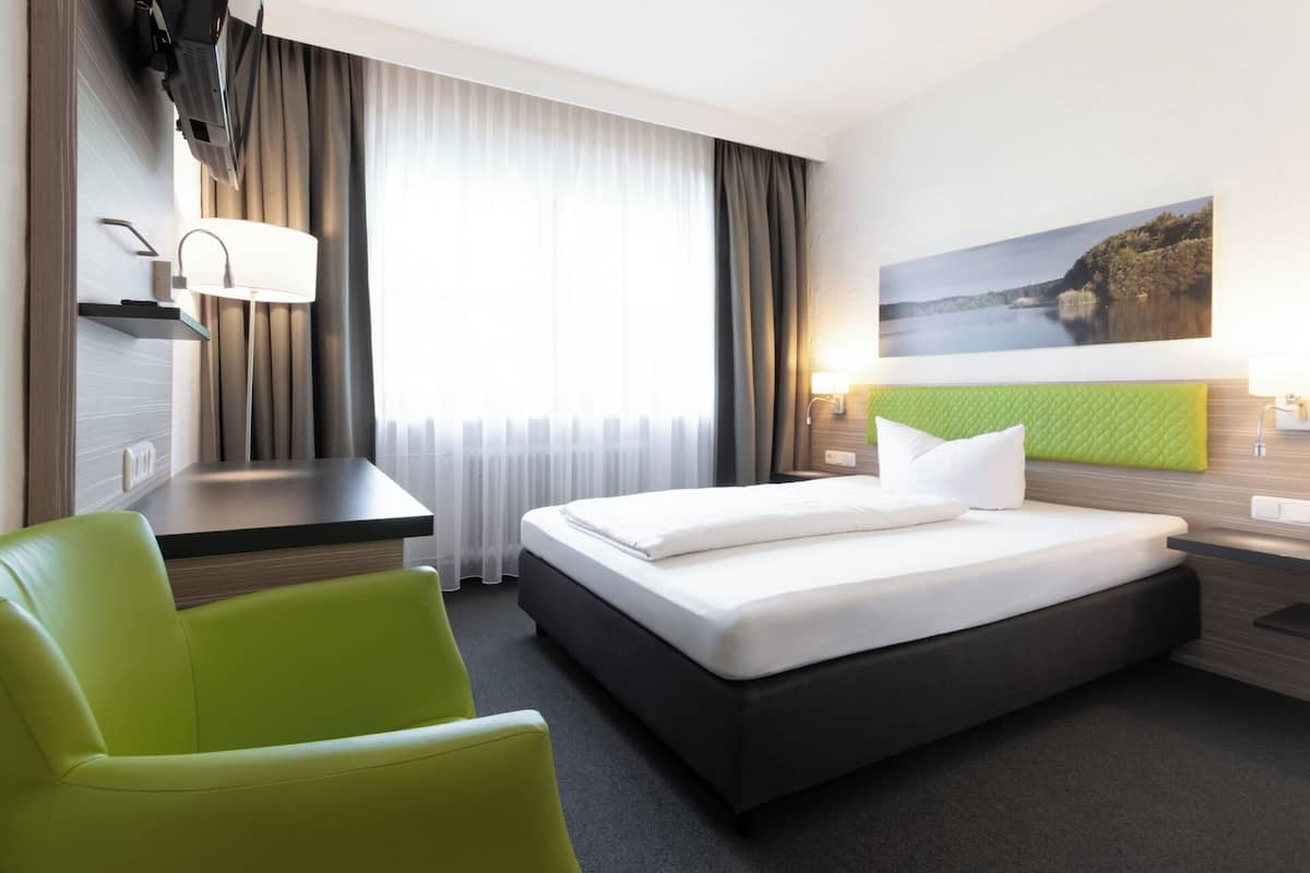 Une chambre d’hôtel avec un lit, un bureau, une chaise, une fenêtre avec des rideaux, un tableau au mur et une lampe.