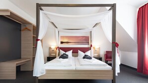 Ein Himmelbett mit weißen Leinen und einem roten Kopfteil, ein Nachttisch mit Lampe, ein roter Sessel und Blick auf einen Sonnenuntergang durch ein Fenster.
