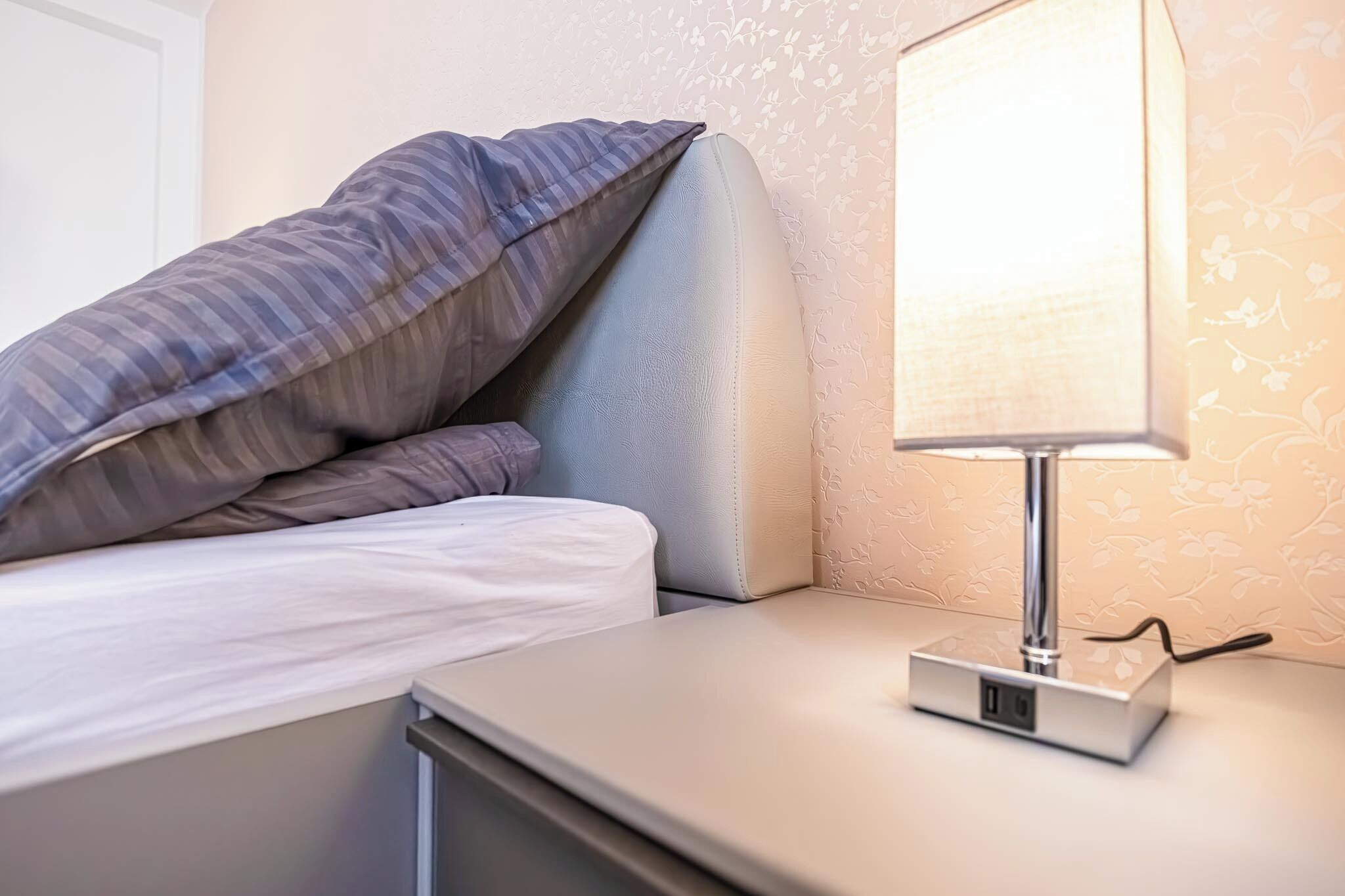 A neatly made bed with a striped pillow and a bedside table with a lamp and a power strip.