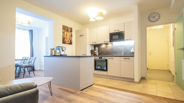 Een compacte keuken met een centraal eiland, een magnetron en een oven. Een eethoek met een tafel en stoelen is zichtbaar door een open doorgang.