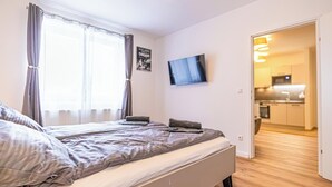 Een slaapkamer met een bed, gordijnen, een flatscreen-tv en uitzicht op de keuken.