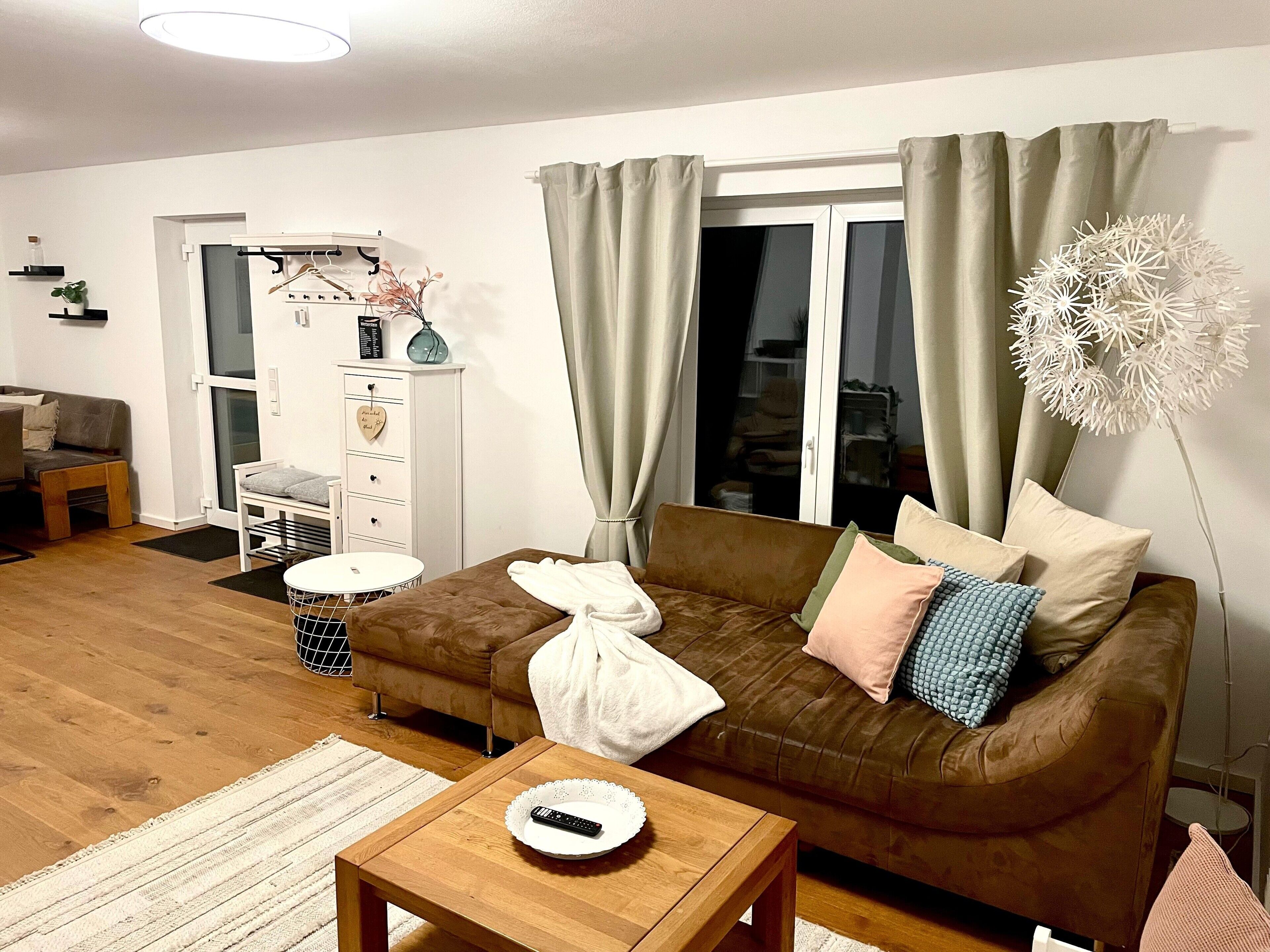 Een woonkamer met een bruine bank, een witte tafel en een groot raam met gordijnen.