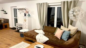 Een woonkamer met een bruine bank, een witte tafel en een groot raam met gordijnen.