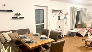 Un coin repas avec une table en bois dressée pour quatre personnes, un canapé avec des coussins, une petite plante et un meuble blanc muni d’une étagère suspendue.