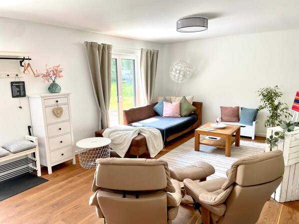 Ein modernes Zimmer mit Bett, Sofa, Sesseln, einem kleinen Tisch und einem Fenster mit Vorhängen.
