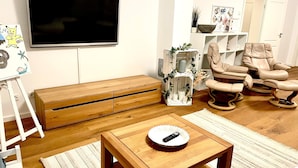 Un salon comprenant un téléviseur, des meubles en bois, une console blanche, un fauteuil en cuir beige et un meuble de rangement blanc agrémenté d’objets décoratifs.