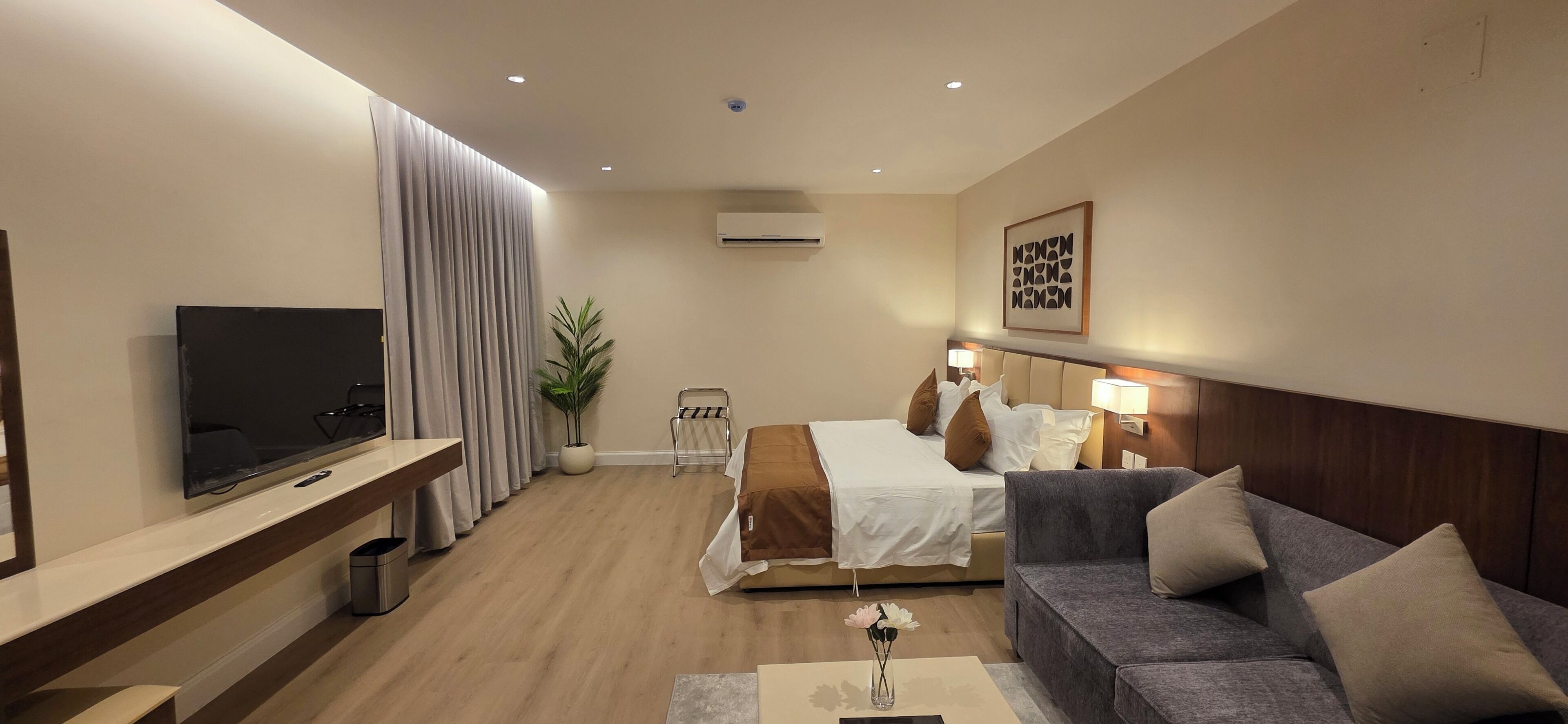 A modern hotel room with a flat-screen TV, a bed with white and brown bedding, a grey sofa, and a small table with a vase.