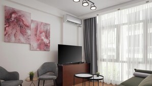 Ein modernes Wohnzimmer mit Fernseher, zwei grauen Sesseln, einem runden Couchtisch und einem grünen Sofa. An der Wand hängen zwei abstrakte Gemälde und ein großes Fenster mit durchsichtigen Vorhängen.