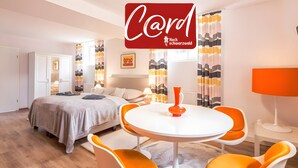Ein modernes Hotelzimmer mit einem Bett, einem runden Esstisch mit orangefarbenen Stühlen, einem weißen Kleiderschrank und einem großen Fenster mit Vorhängen.
