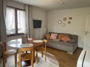 A living room with a round wooden table, two chairs, a grey sofa with cushions, a radiator, and wall decorations.