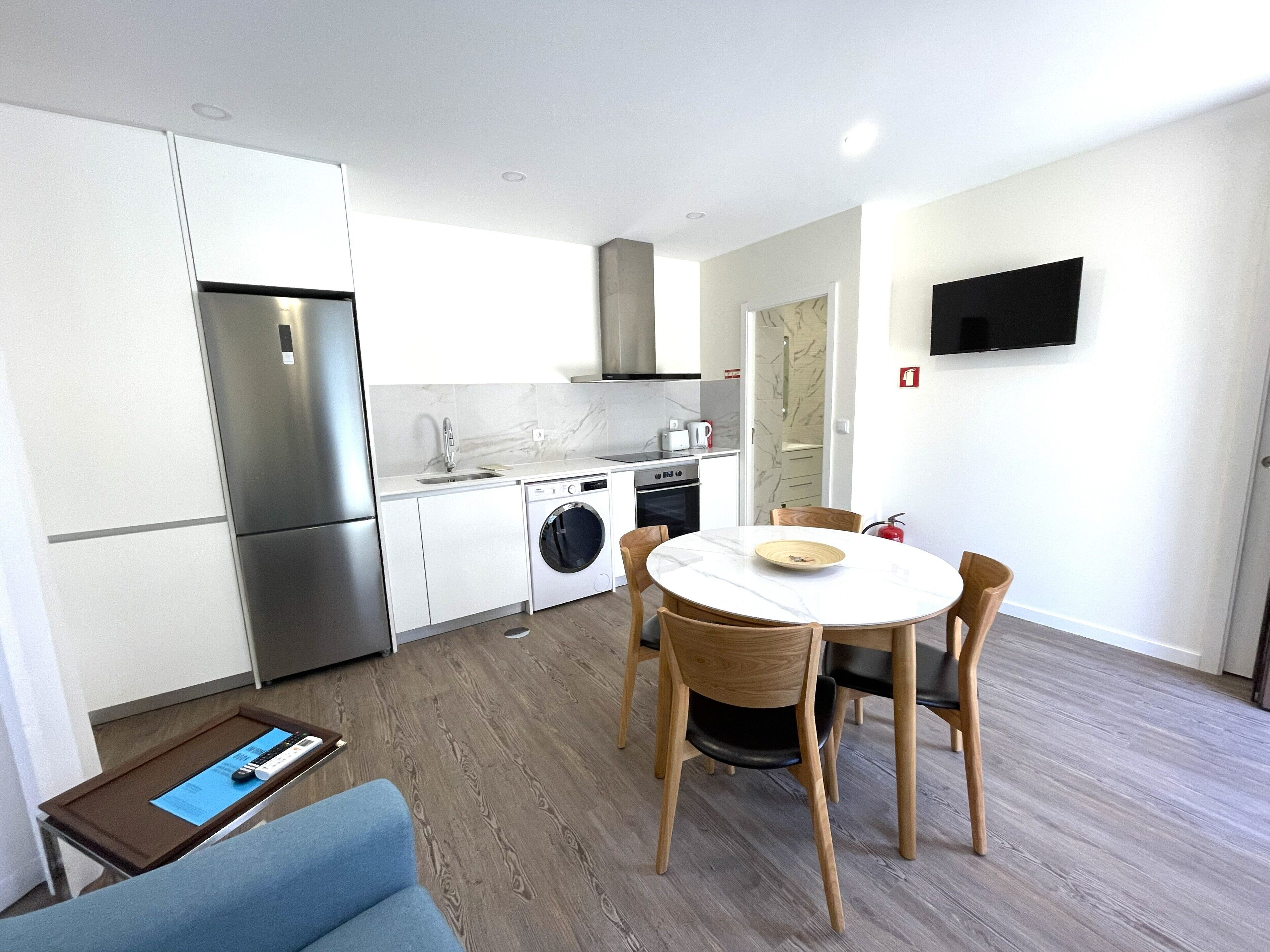 A modern kitchen with stainless steel appliances, a washing machine, and a dining area with a round table and wooden chairs.