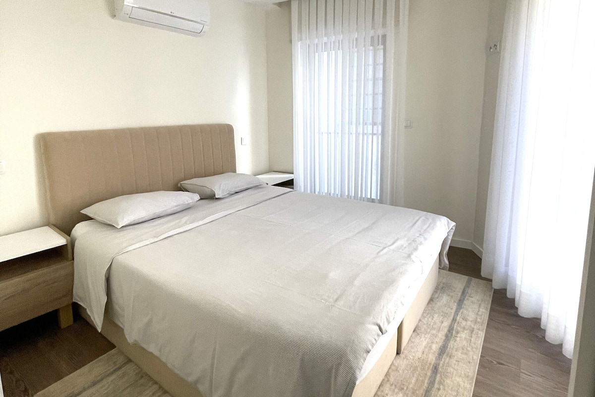 A modern hotel room with a large bed, a bedside table, an air conditioning unit, and a window with curtains.