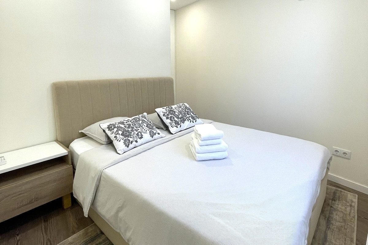 A neatly made bed with white linens and two decorative pillows, a bedside table with a lamp, and an air conditioning unit on the wall.