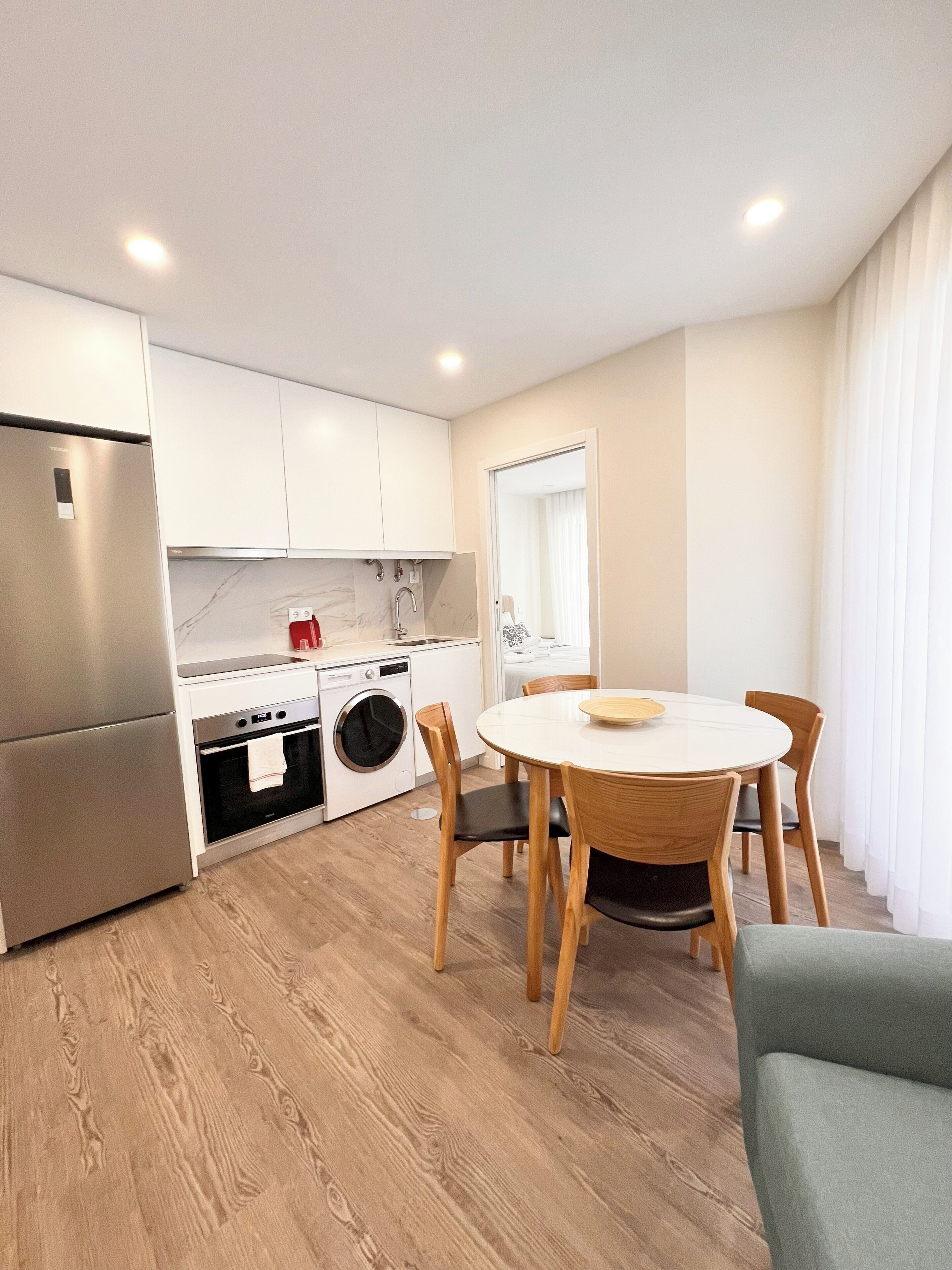 Una cocina moderna con armarios blancos, un refrigerador de acero inoxidable, un horno integrado y una lavadora. Hay una mesa redonda de comedor con sillas de madera y un sofá de color claro.