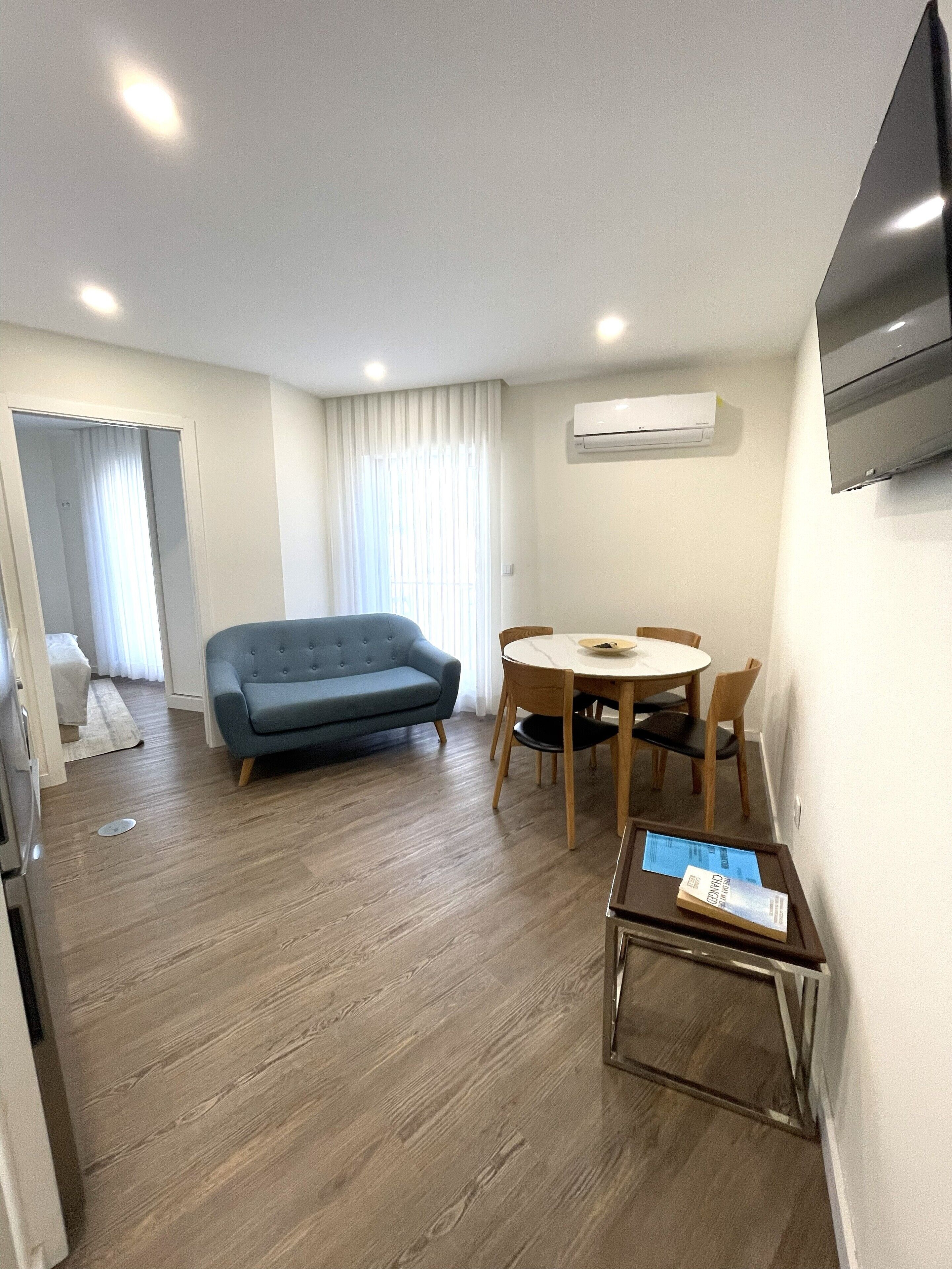 A modern living room with a blue sofa, wooden dining table, and chairs. There is a flat-screen TV mounted on the wall and an air conditioning unit above the TV.