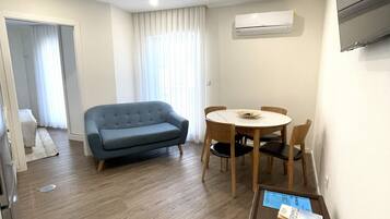 Una sala moderna con un sofá azul, una mesa de comedor de madera y sillas. Hay un televisor de pantalla plana montado en la pared y un equipo de aire acondicionado encima.