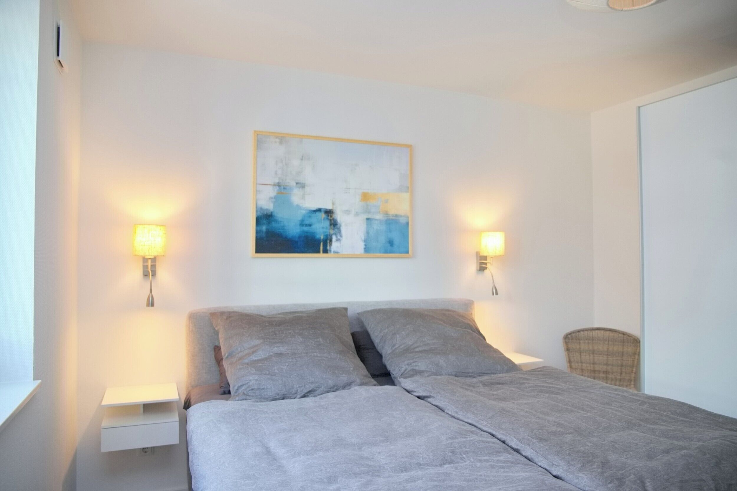 Ein Schlafzimmer mit einem Bett, zwei Nachttischlampen, einem Wandbild im abstrakten Stil und einem Sessel.