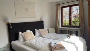 A neatly made bed with white linens and two beige pillows, a bedside table with a lamp, and a window with curtains. - 'Sandvoss' 3-Room Apartment by Gästehaus Sandvoss, Near Lake, Titisee (Titisee-Neustadt)