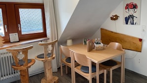 Ett rum med ett träbord och stolar, ett fönster med gardiner, ett element och ett konstverk som hänger på väggen.