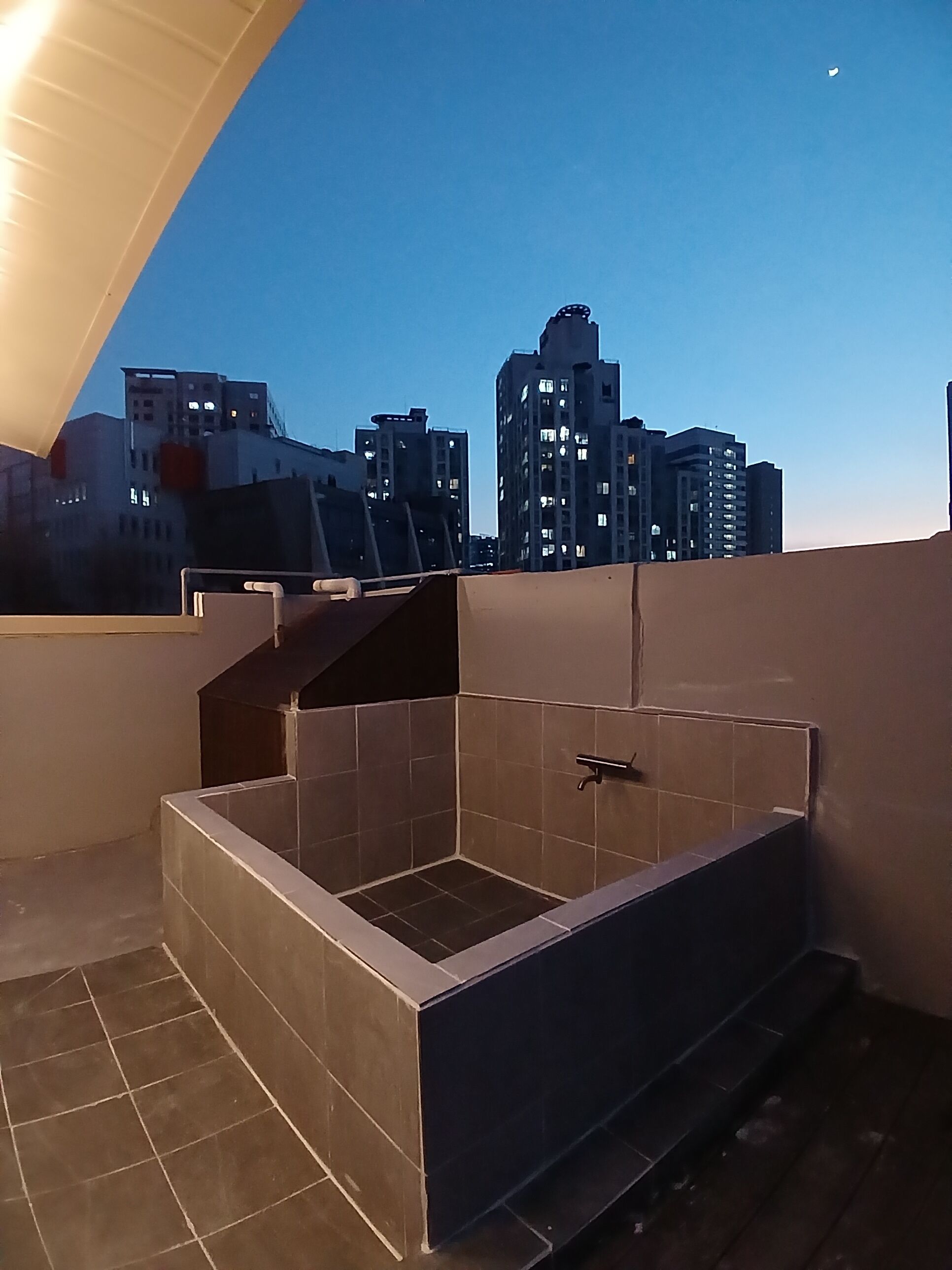 Une terrasse sur le toit avec une baignoire carrelée, une rambarde en métal et une vue sur les toits de la ville en arrière-plan.
