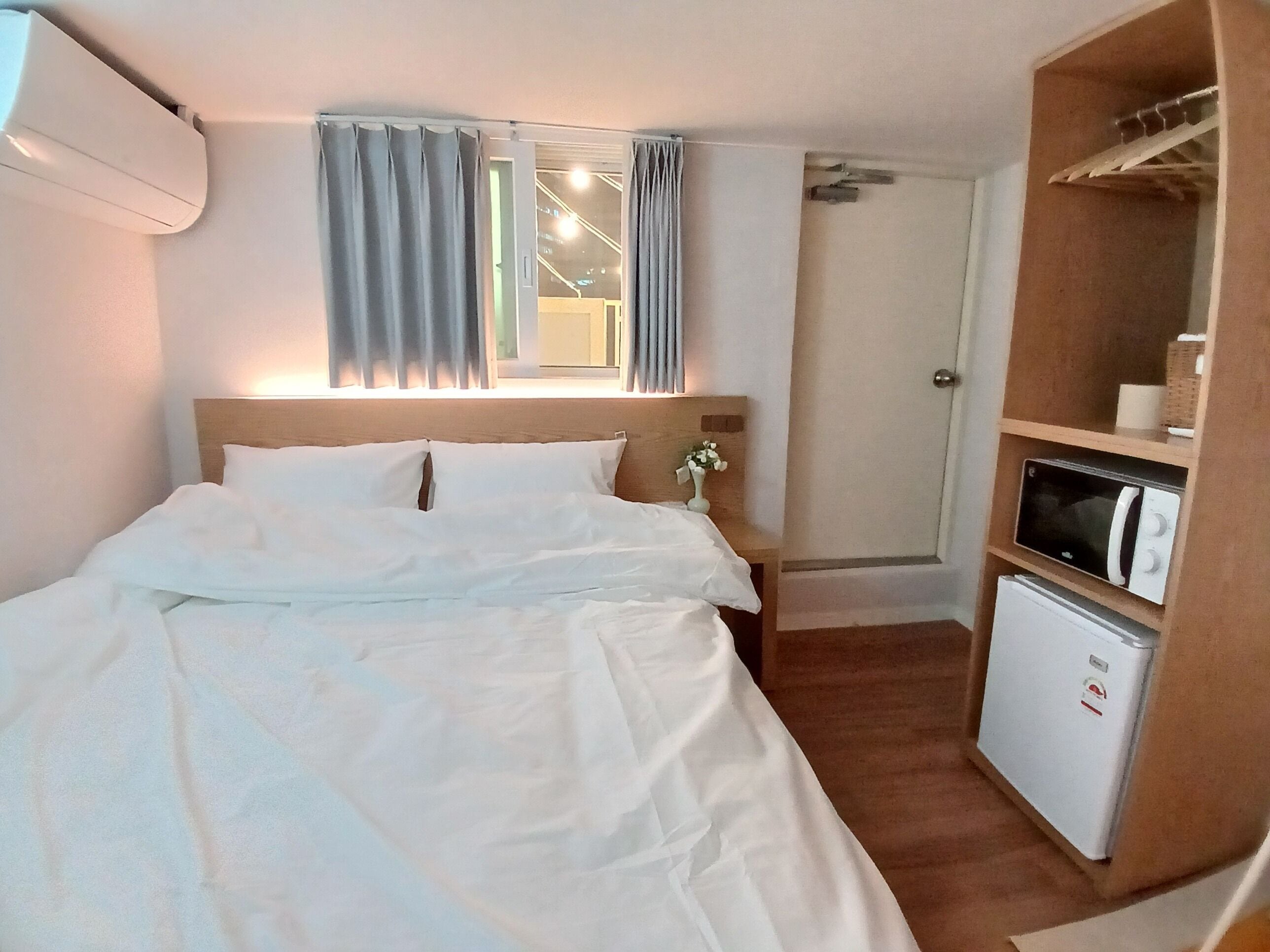 Habitación de hotel con cama, mesita de noche, ventana con cortinas, microondas integrado y un refrigerador pequeño.