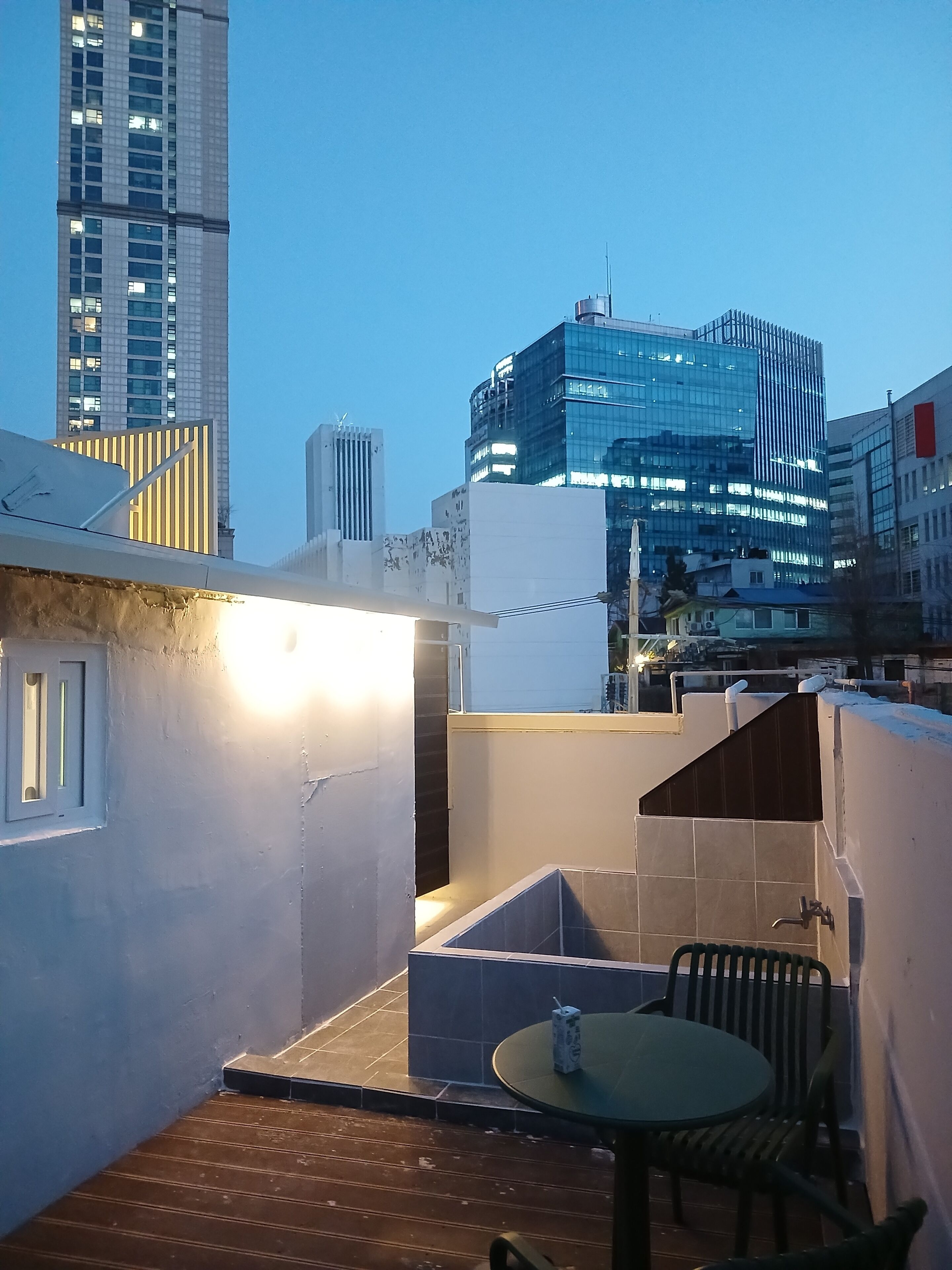 A rooftop terrace with a table and chairs, a view of modern buildings, and a clear sky.