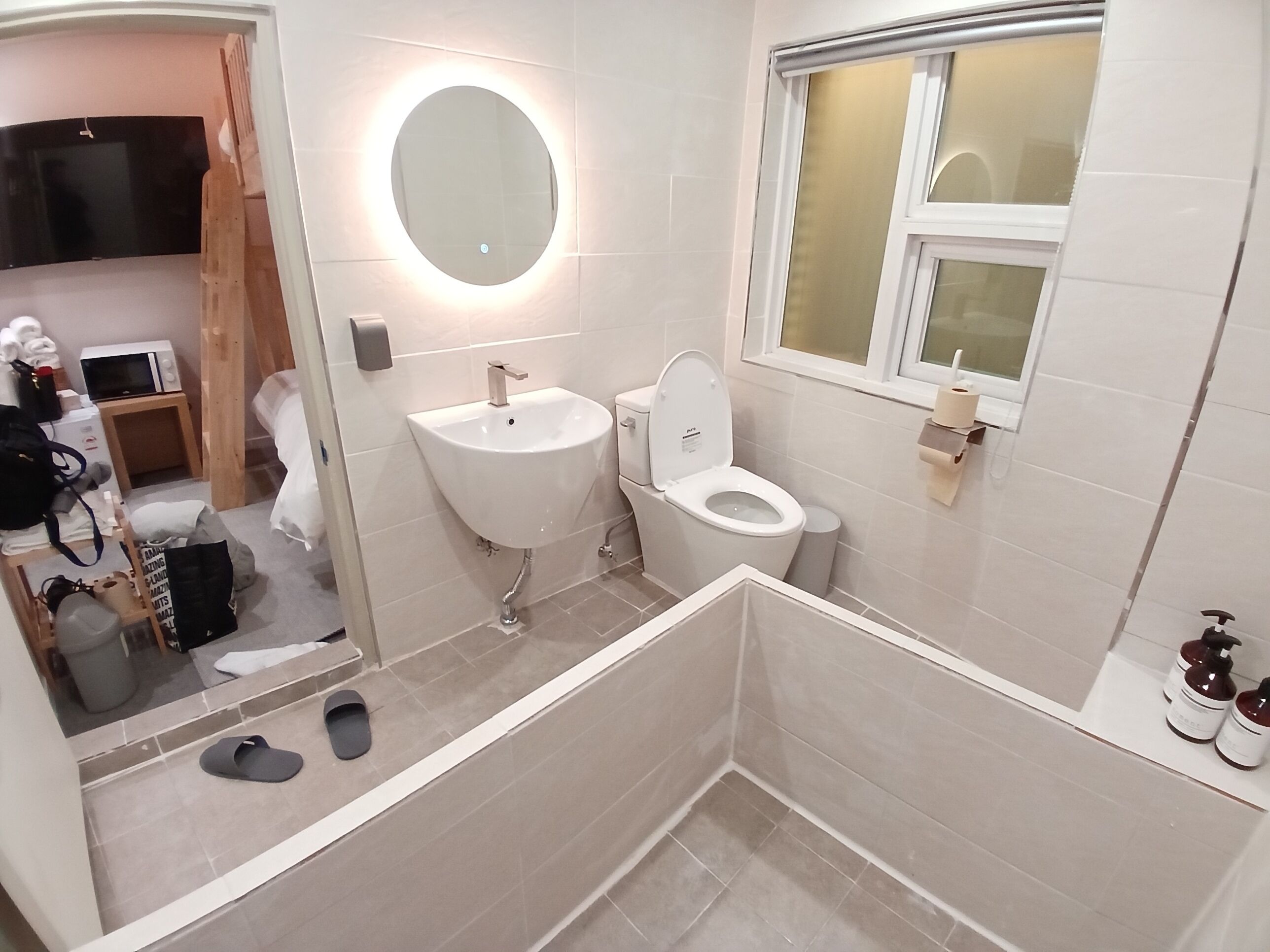 Et moderne badeværelse med vask, toilet og et spejl med lys. Der er et vindue og en lille hylde med toiletartikler.