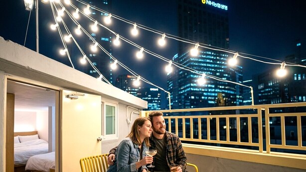 Una pareja sentada en un balcón con luces colgantes, una botella de licor y un bol de aperitivos.