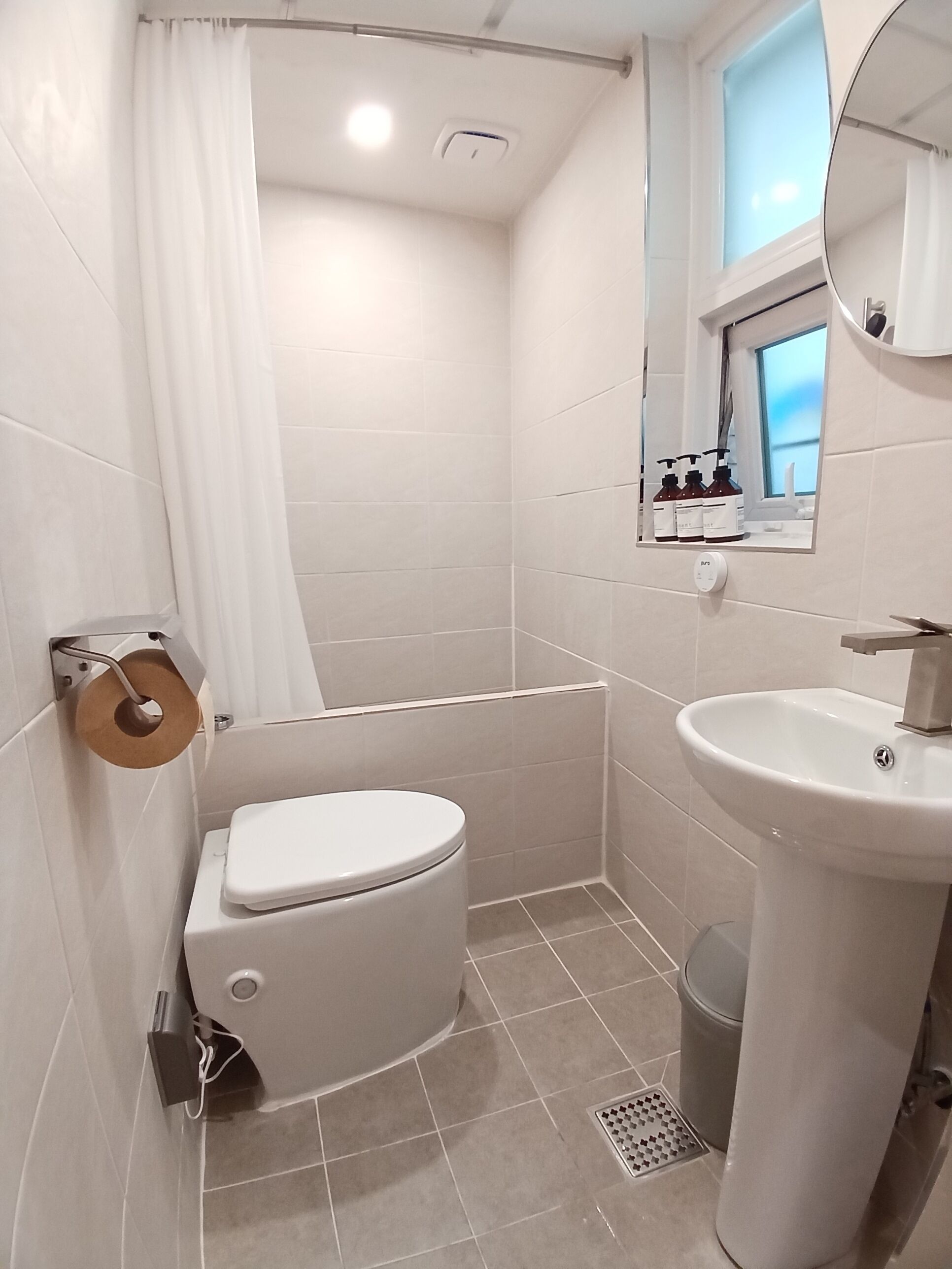 A bathroom with a toilet, sink, shower, and a window with toiletries.