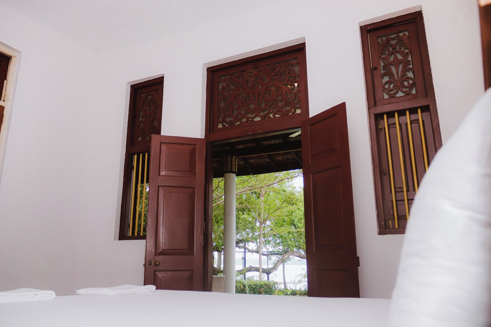 A room with a bed, a window with wooden shutters, and a view of greenery outside.