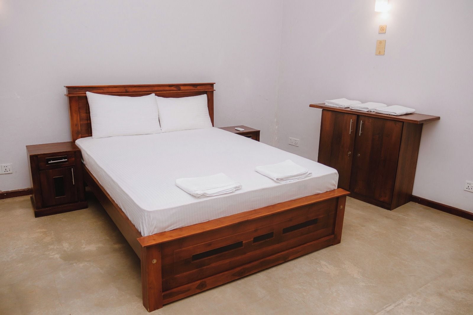 Ein ordentlich bezogenes Bett mit weißen Textilien und zwei Kissen, ein Nachttisch aus Holz mit Schublade, ein Holzschrank und ein kleiner Tisch mit gefalteten Handtüchern.