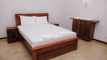 Ein ordentlich bezogenes Bett mit weißen Textilien und zwei Kissen, ein Nachttisch aus Holz mit Schublade, ein Holzschrank und ein kleiner Tisch mit gefalteten Handtüchern.