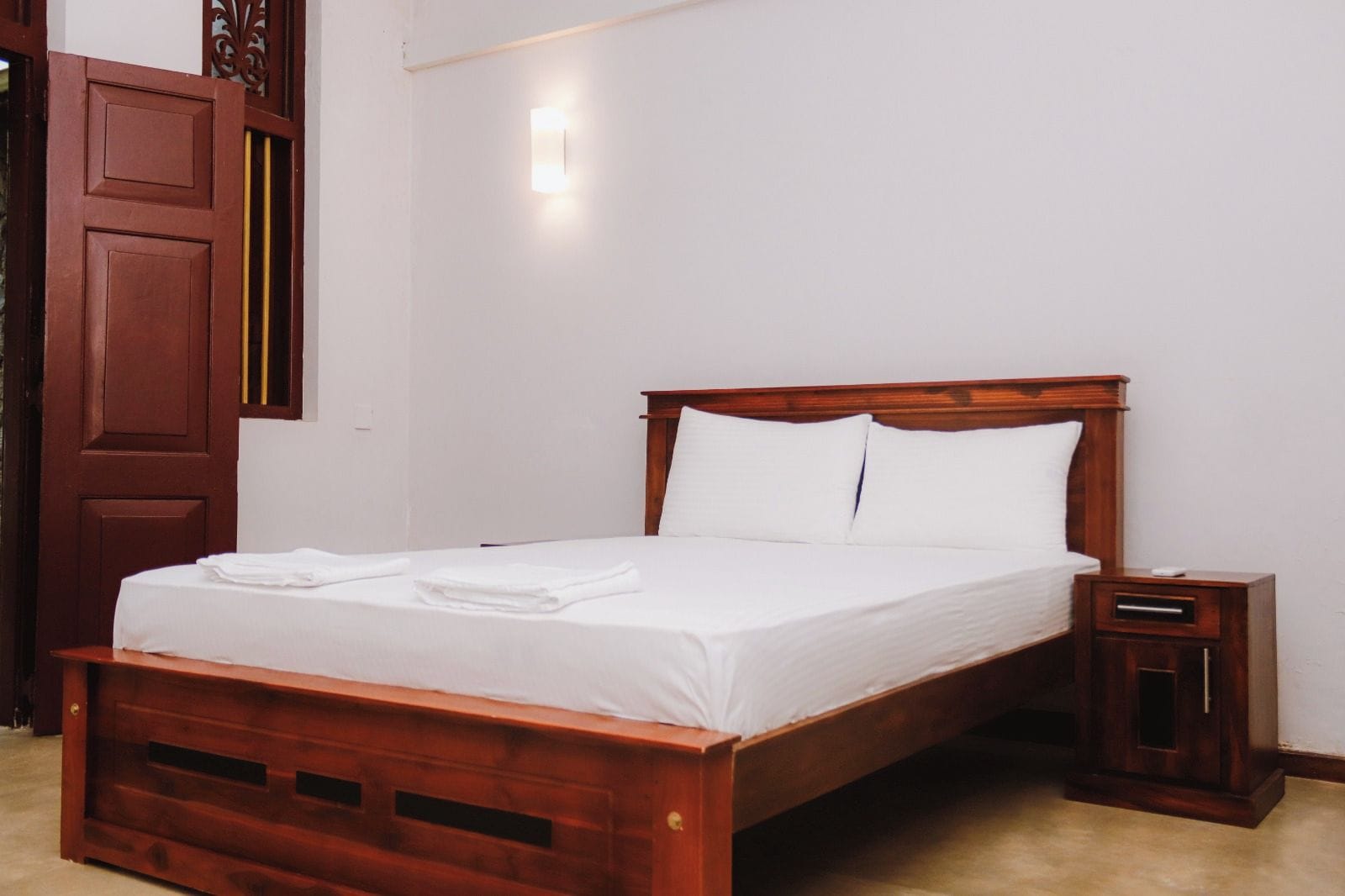 A neatly made bed with white linens and pillows, a wooden headboard, and a small bedside table with a drawer.