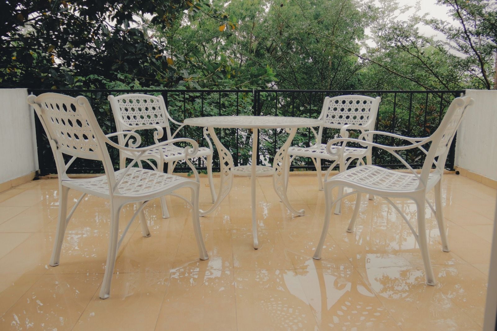 A set of four white metal chairs and a table arranged on a tiled patio with a black metal railing in the background.