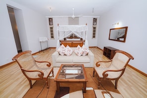 A living room with a sofa, two armchairs, a glass coffee table, a ceiling fan, and a bed with a canopy.
