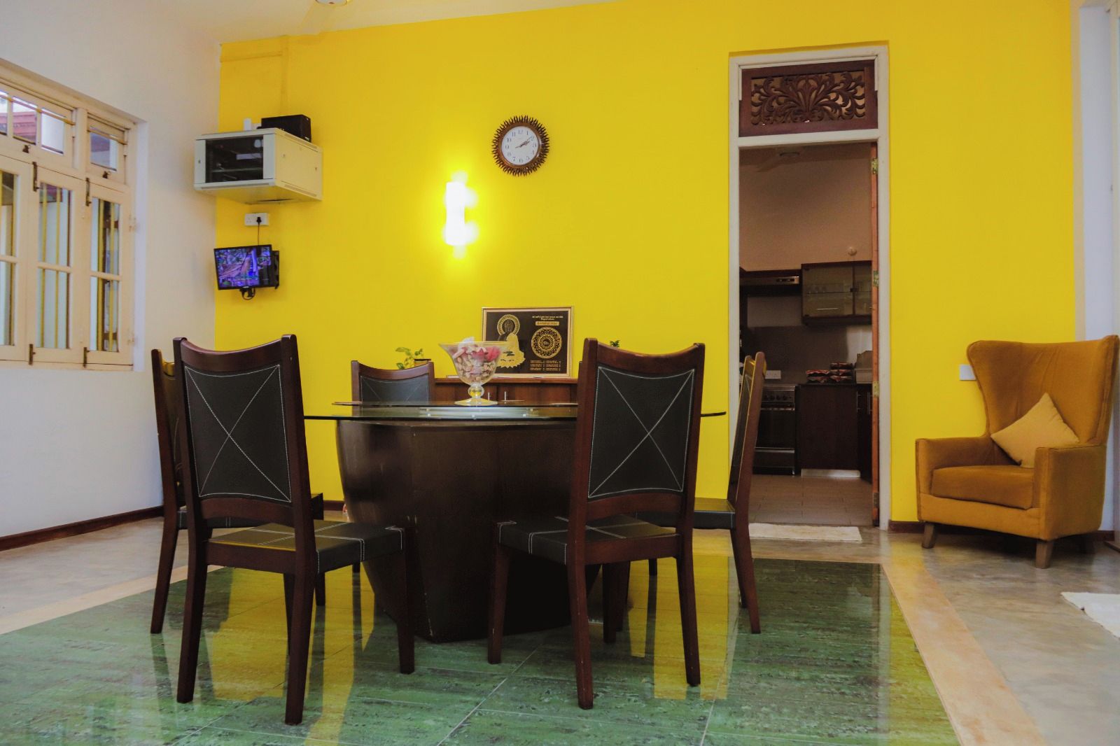 Un'area pranzo con un tavolo rotondo e quattro sedie, una sveglia appesa al muro e un televisore su una mensola.