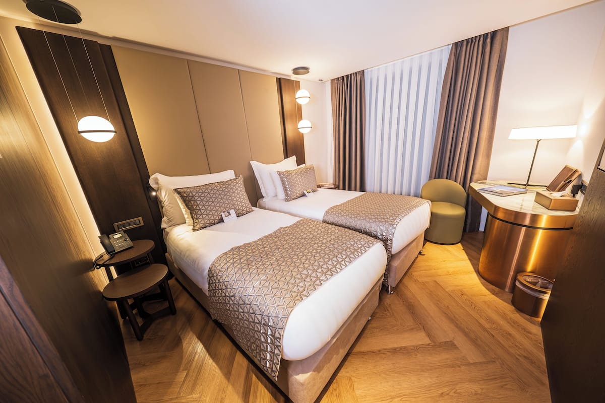 A hotel room with two beds, a desk, a chair, a lamp, and a trash bin.