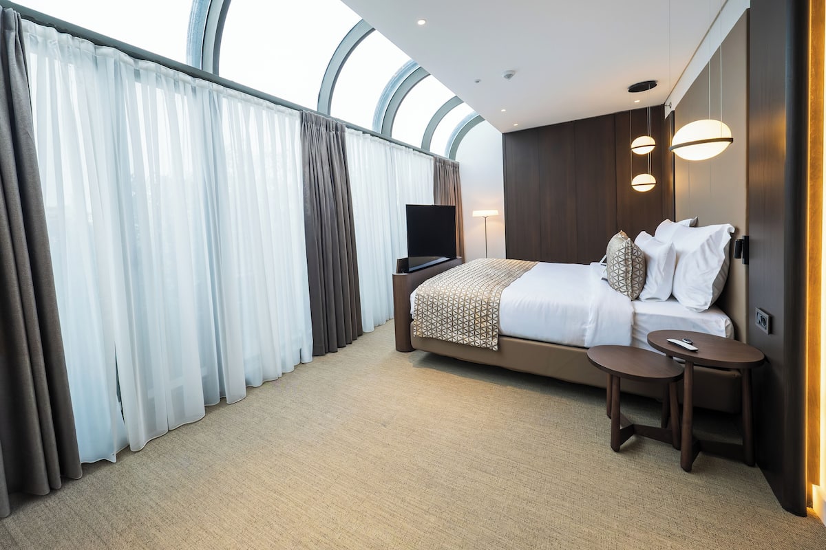 A modern hotel room with a large bed, a TV, a bedside table, and a view through a large window.