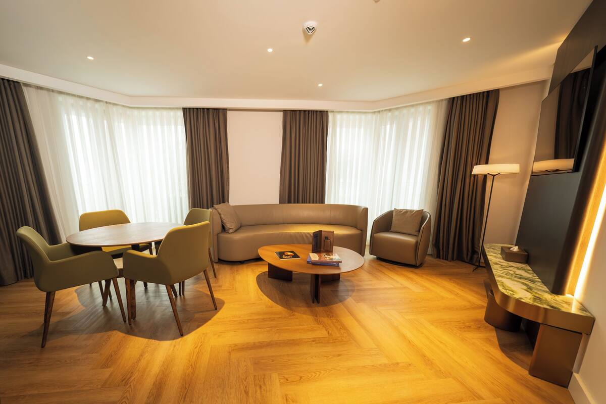 A modern living room with a round coffee table, a sofa, and two armchairs. There is a dining table with chairs, and the room has large windows with curtains.