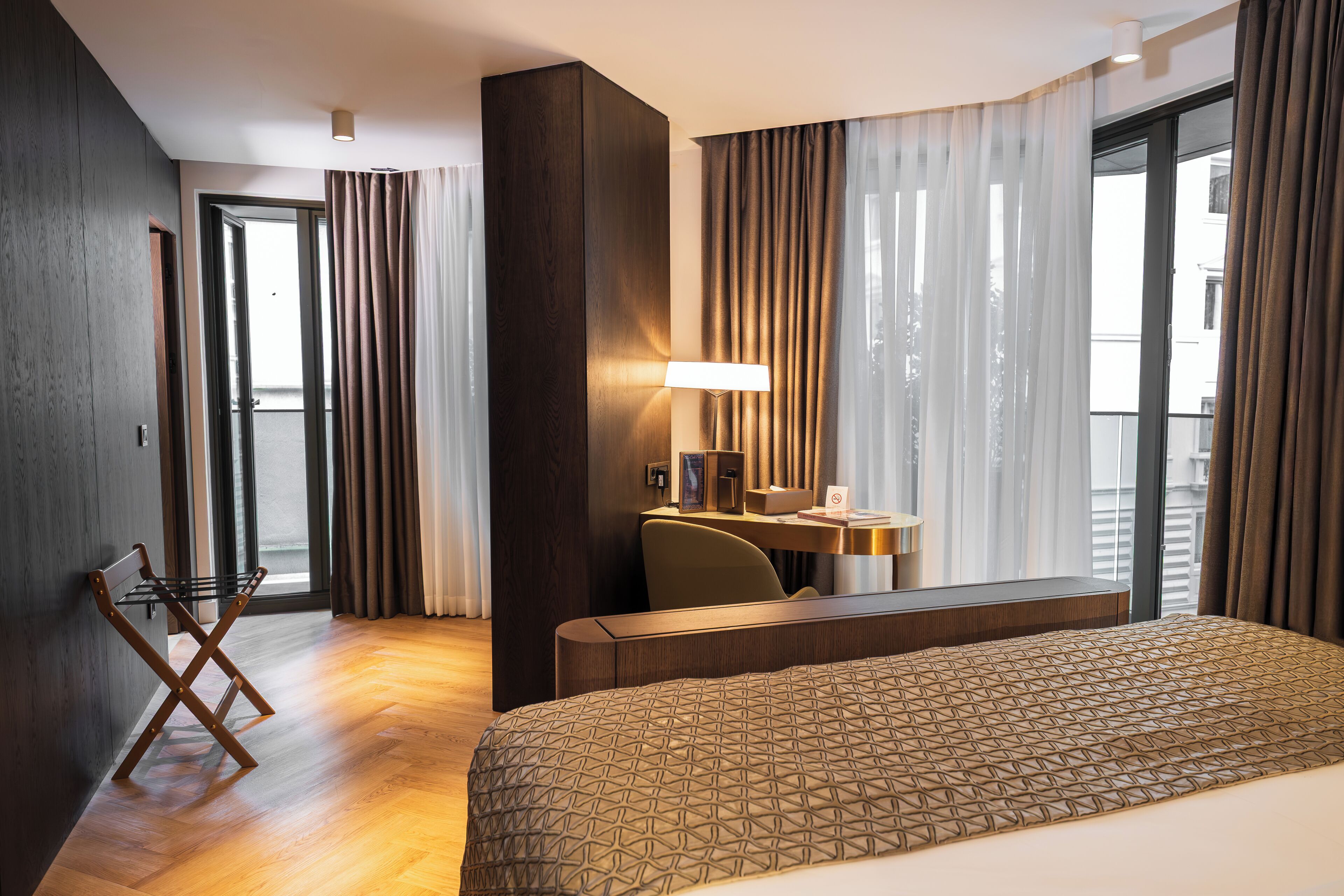 A hotel room with a large bed, a desk with a chair, a standing lamp, and a view of the outside through large windows.