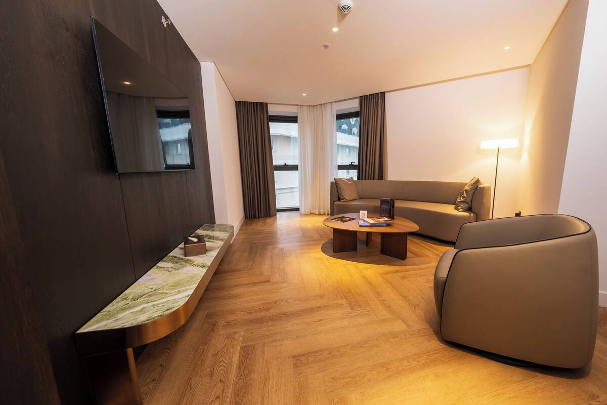 A modern hotel room with a wooden floor, a sofa, a coffee table, a TV mounted on the wall, and large windows with curtains.