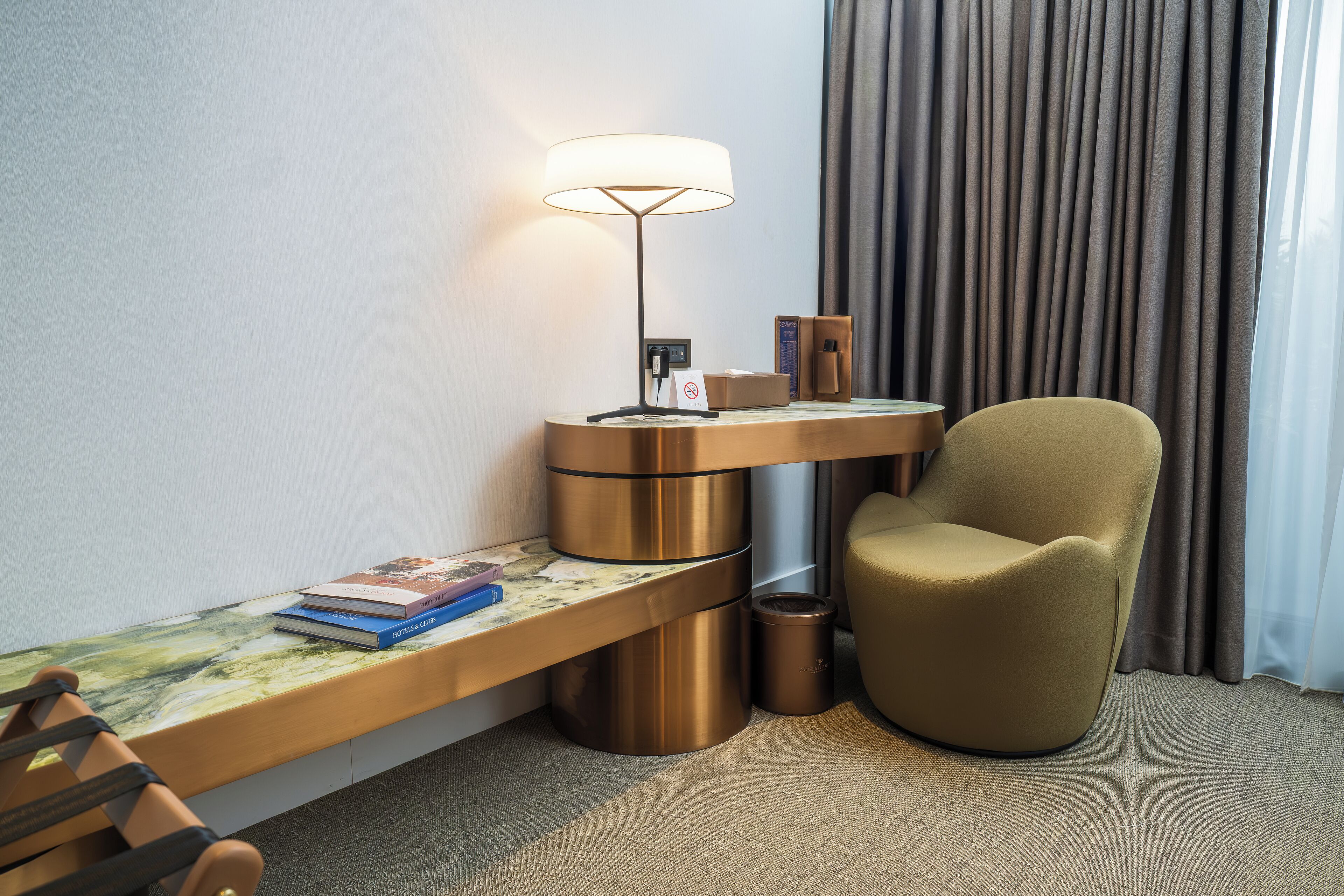 A modern hotel room with a desk, chair, lamp, and a view of the outside through curtains.