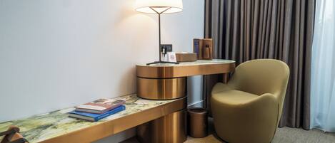 A modern hotel room with a desk, chair, lamp, and a view of the outside through curtains.