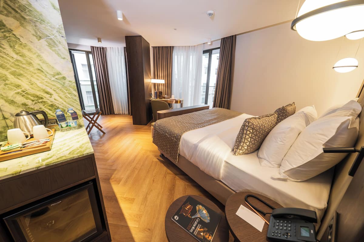 A hotel room with a bed, bedside table, lamp, and a view of the city through large windows.