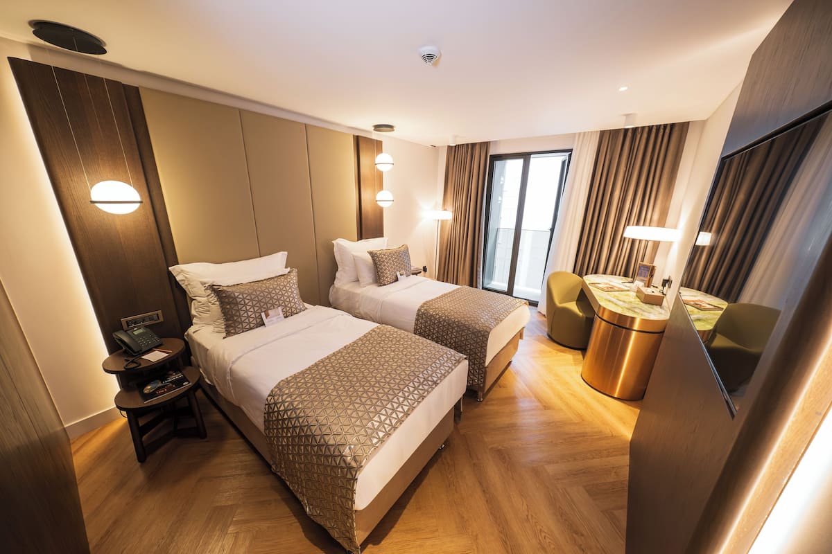 A hotel room with two beds, a small table with a lamp, a chair, and a large window.