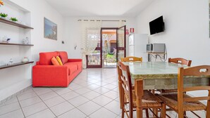 Een woonkamer met een rode bank, een eettafel met stoelen, een televisie en een keukenhoek met een magnetron.