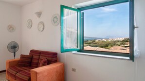 Une pièce comprenant un canapé, un ventilateur sur pied, une fenêtre donnant sur la côte et des assiettes décoratives accrochées au mur.